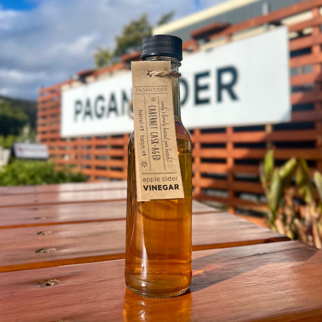 A bottle of Pagan Cider Vinegar with a label that includes the product name and 'AGED IN OAK CABERNET SAUVIGNON WINE BARRELS 10 MONTHS' text, placed on a wooden surface with a building in the background.