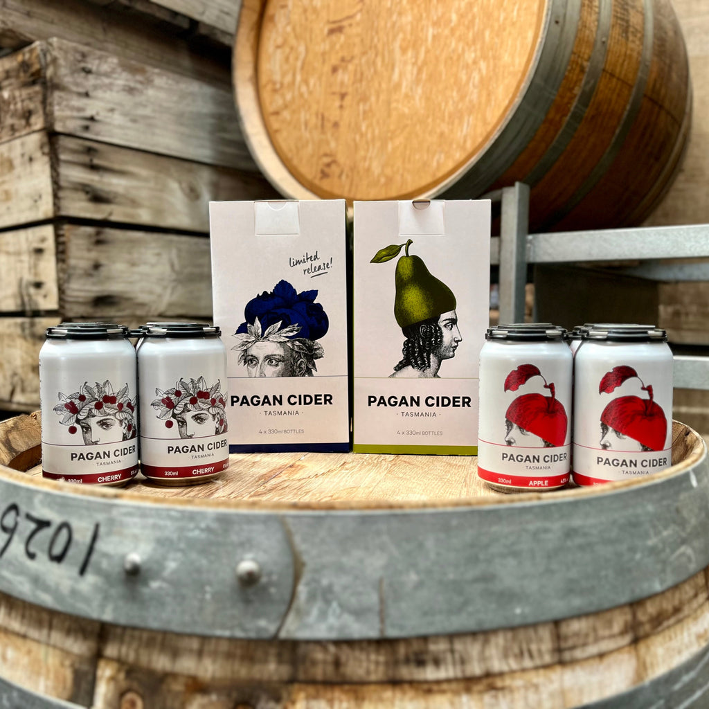 A selection of Pagan Cider products displayed on a wooden barrel, including cans and bottles with labels showing different flavors such as cherry, blueberry, pear, and apple.
