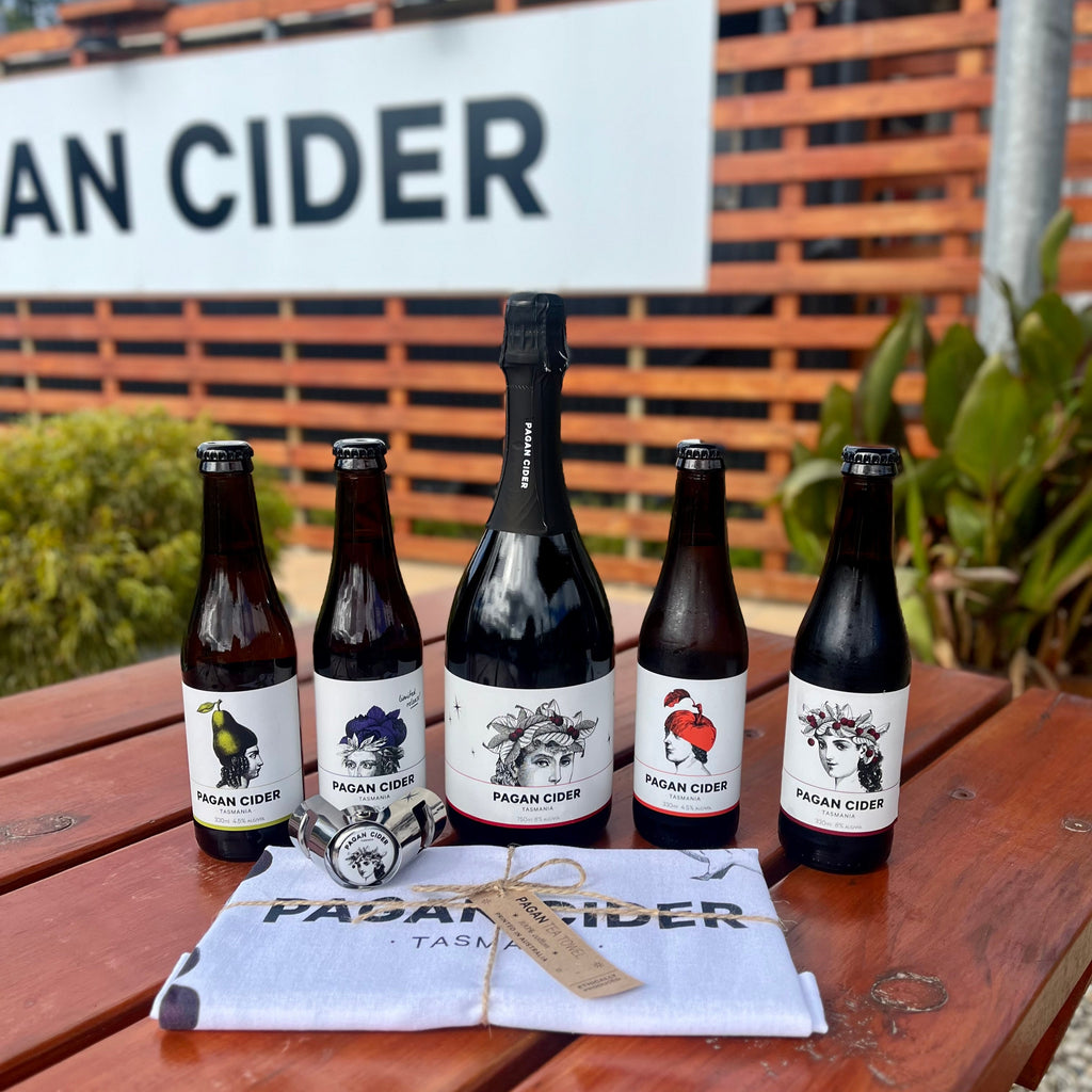 A set of bottles of various ciders from Pagan Cider, including a bottle of Cherry Sparkle, arranged on a table with a 'PAGAN CIDER' sign in the background. There's also a tea towel with the brand's logo on it.