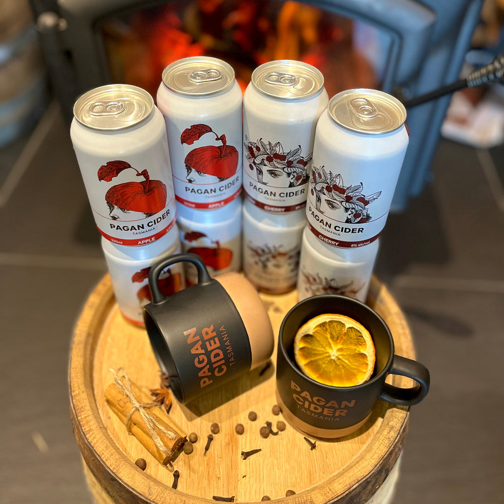 A set of Pagan cider cans and mulled cider in a mug, with spices and a lemon slice, placed on a wooden round table in front of a log fire.